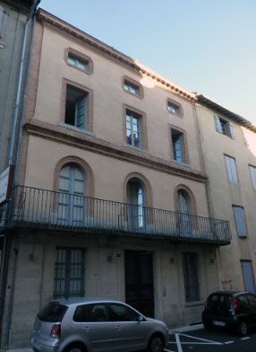 Immeuble beige de trois étages avec fenêtres cintrées, balcon et voitures garées devant.