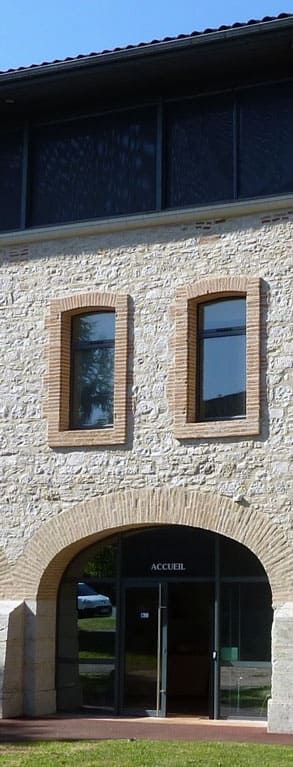 Bâtiment en pierre avec entrée et fenêtres cintrées. La lumière du soleil se reflète sur les vitres.