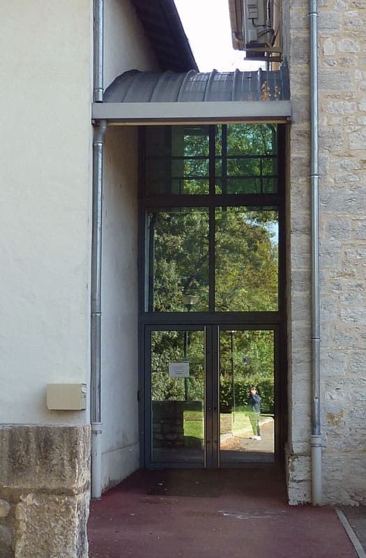 Porte extérieure vitrée, encadrée par des murs couleur crème et en pierre, donnant sur un espace vert.
