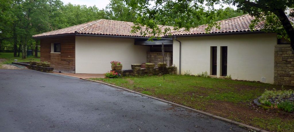 Bâtiment bas avec toit texturé, murs beiges et petite pelouse verte devant. Allée asphaltée.