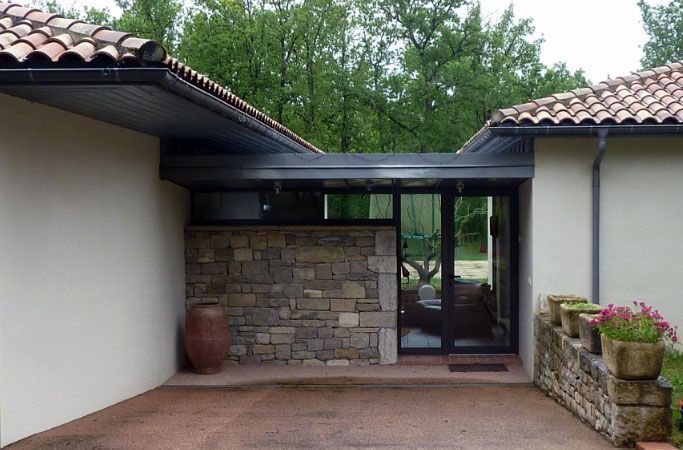 Extérieur d'une maison avec porte vitrée, mur en pierre, toit en tuiles de terre cuite et végétation environnante.