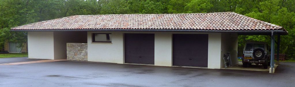 Un petit bâtiment avec deux portes de garage et un abri voiture. Une voiture est garée sous l'abri. Le bâtiment a un toit en tuiles.