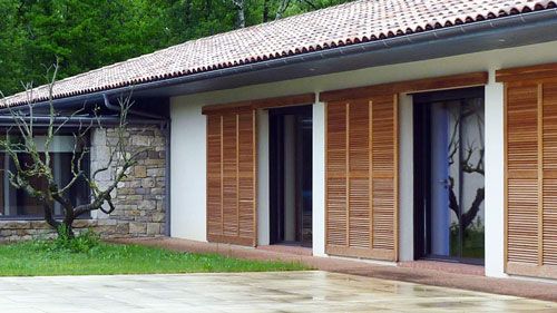 Extérieur d'une maison aux volets bruns, mur en pierre et toit de tuiles rouges. Jardin arboré et pelouse.