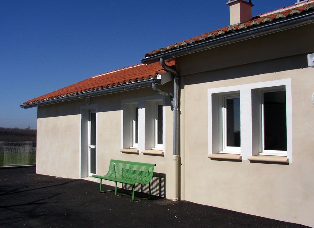 Bâtiment en stuc beige avec toit de tuiles rouges, fenêtres à encadrement blanc, banc vert. Ciel bleu.
