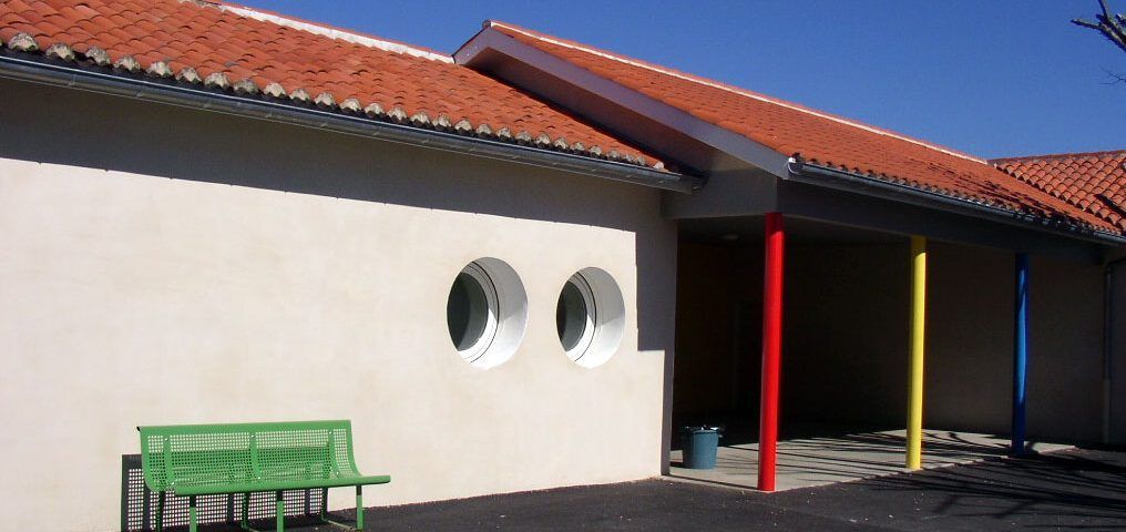 Bâtiment au toit de tuiles, aux murs blancs et aux poutres de soutien colorées. Un banc vert est installé devant.