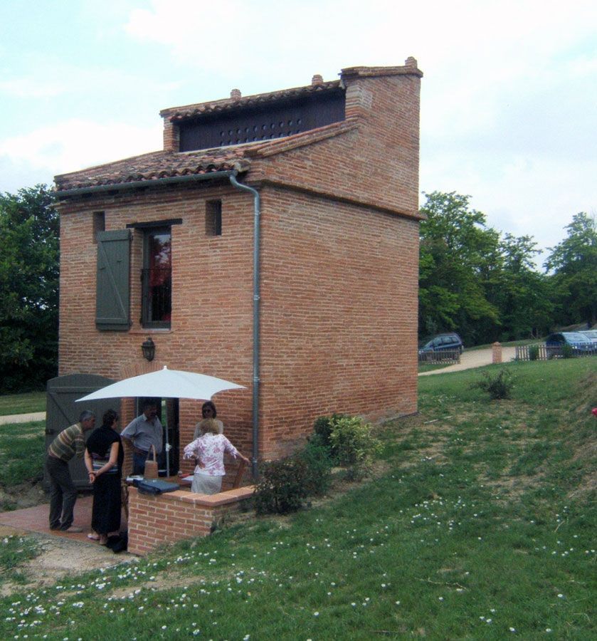 Bâtiment en briques rouges avec des personnes sous un parasol blanc, entouré d'herbe verte et d'arbres.