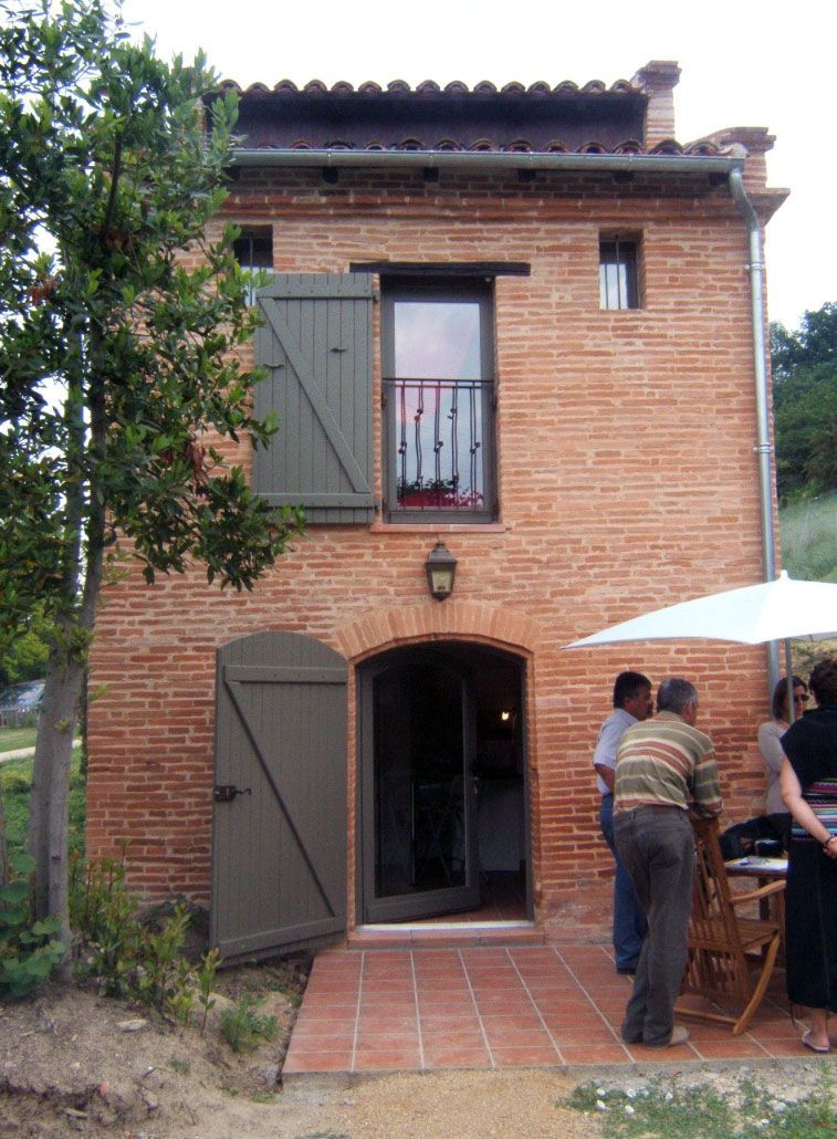Bâtiment en briques aux volets verts et à la porte ouverte, des gens rassemblés à l'extérieur sur une terrasse.