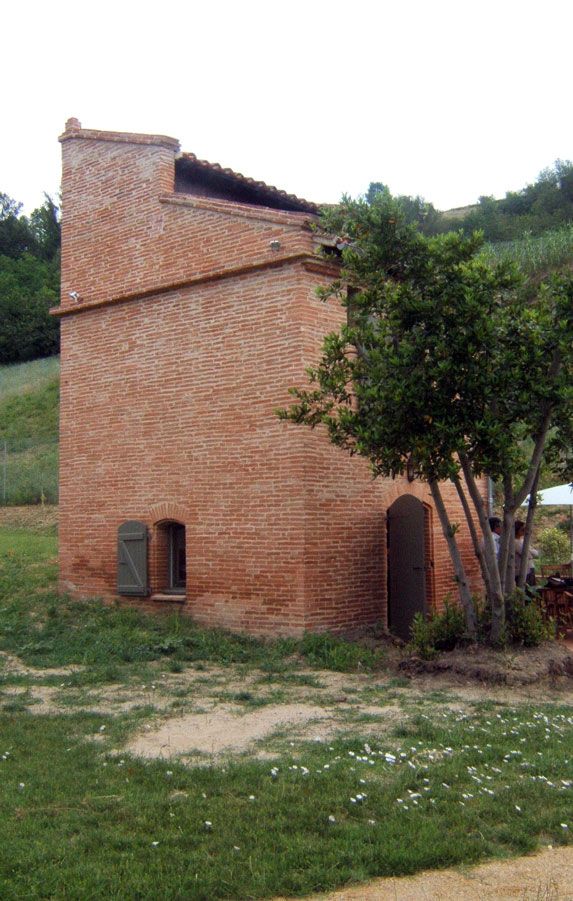 Bâtiment en briques avec toit en pente, petite fenêtre à volets et porte en bois, arbre à droite, espace vert herbeux.