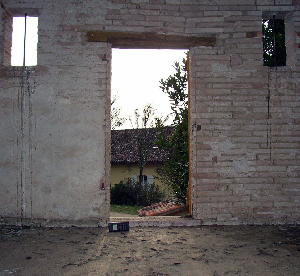 Vue intérieure d'un mur en briques et plâtre avec une porte ouverte. Un bâtiment est visible au loin.