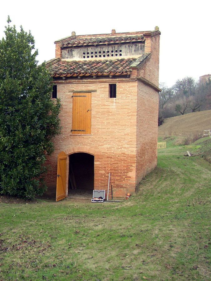 Pigeonnier en briques avec porte ouverte et volets, dans un champ herbeux avec un arbre.