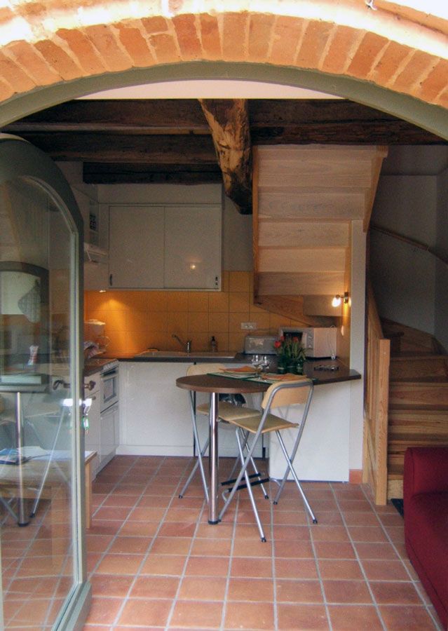 Une porte cintrée en briques encadre une petite cuisine avec une table et des chaises, un escalier et un sol en carrelage de terre cuite.
