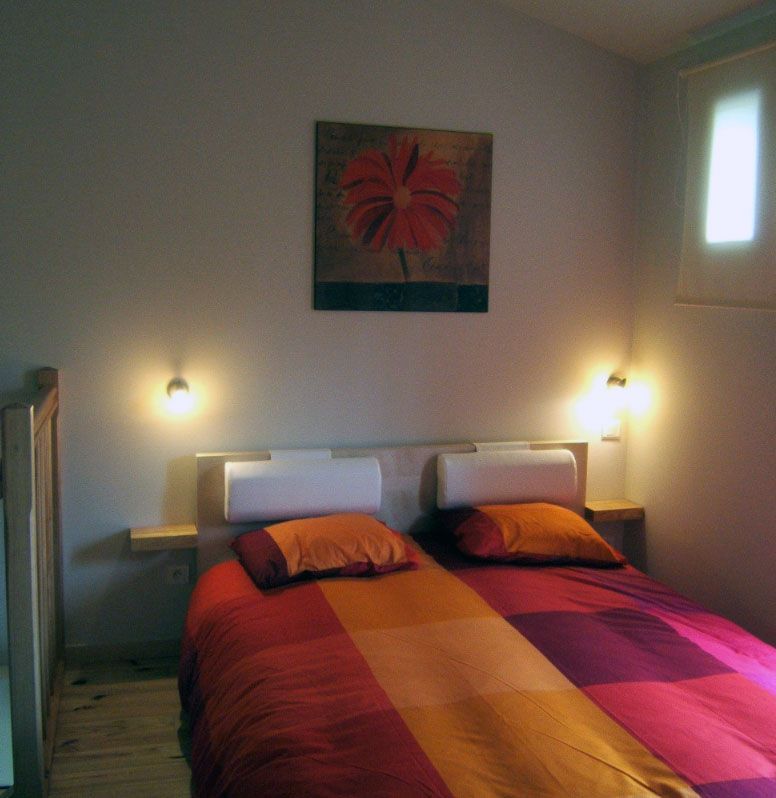 Chambre avec linge de lit rouge et orange, deux oreillers et un tableau représentant une fleur rouge au mur.
