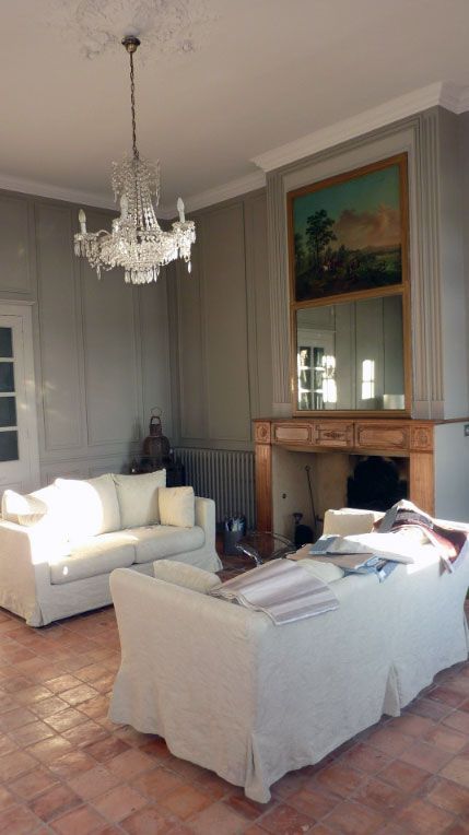 Salon avec deux canapés blancs, un lustre, une cheminée et un tableau au plafond. La lumière du soleil y pénètre abondamment.