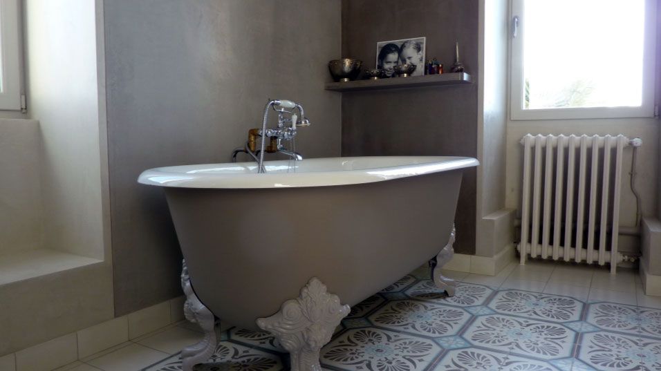 Baignoire sur pieds dans une salle de bains aux murs gris, sol carrelé à motifs et petite étagère avec objets décoratifs.