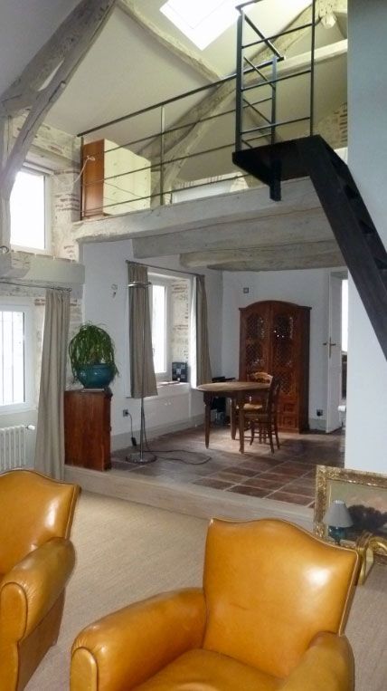 Intérieur avec fauteuils en cuir beige, poutres apparentes et mezzanine avec rampe et escalier noirs.