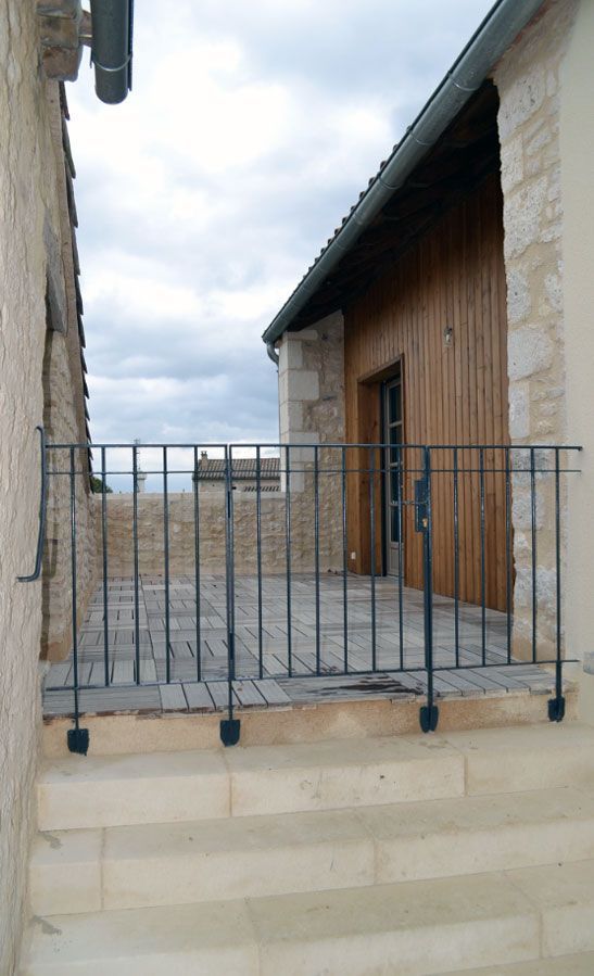 Des marches en pierre mènent à une terrasse dotée d'une balustrade en métal noir. Un bâtiment en bois est visible au-delà de la terrasse.