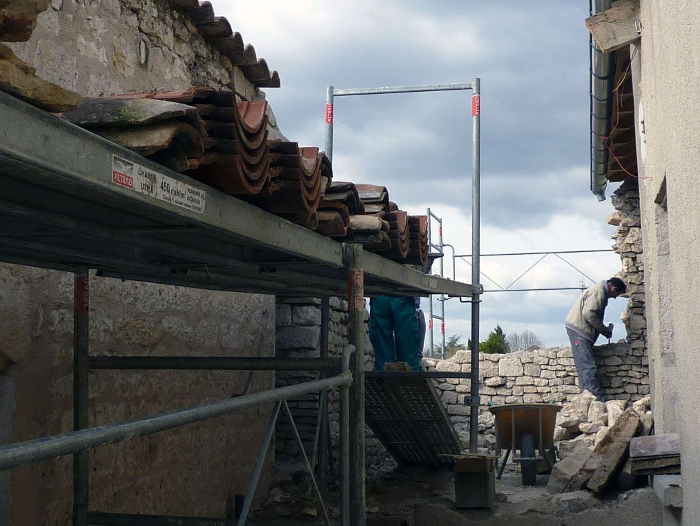 Des ouvriers du bâtiment réparent un immeuble en pierre, utilisant des échafaudages et retirant des tuiles. Ciel couvert.