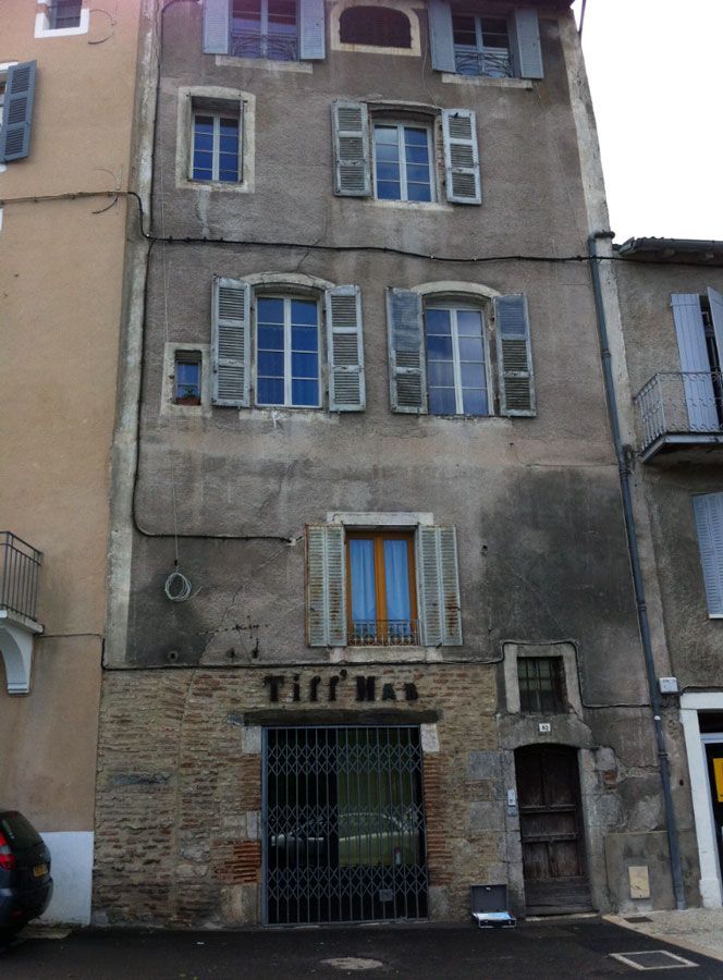 Immeuble en pierre à plusieurs étages avec fenêtres à volets. Panneau Tiff Val au-dessus d'une entrée fermée par un portail.