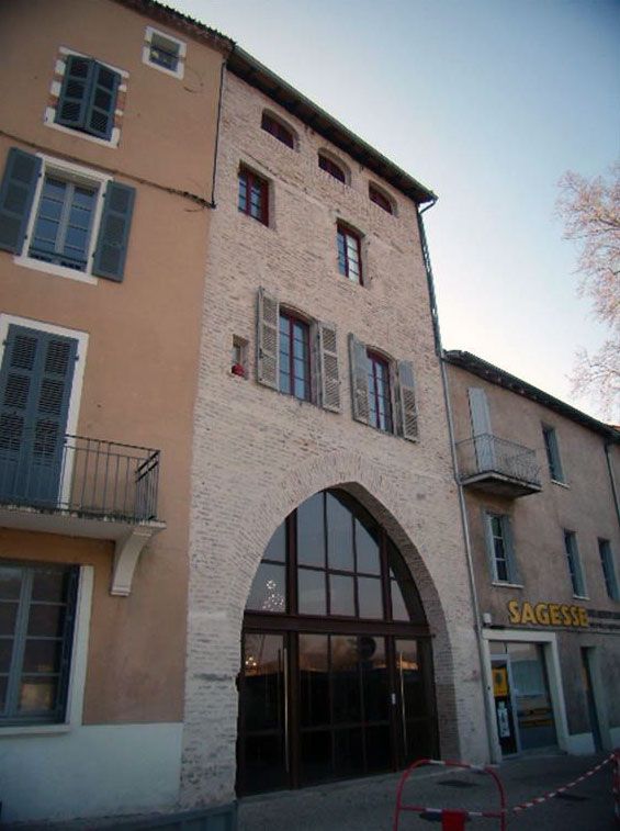 Une arche en pierre menant à un bâtiment, flanquée d'immeubles avec fenêtres et volets, sous un ciel dégagé.