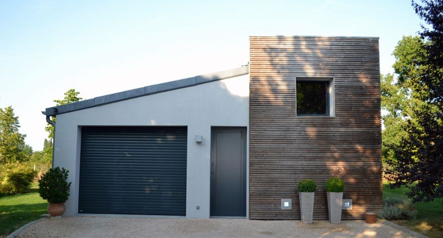 Garage moderne avec une tour revêtue de panneaux de bois. Le garage possède une porte et un volet gris et donne sur une allée de gravier.