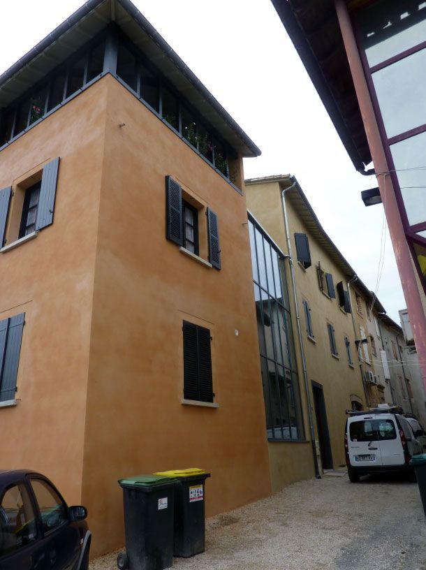 Des immeubles orange et beige dans une rue étroite ; des volets noirs, des fenêtres en verre, une camionnette blanche et des poubelles.