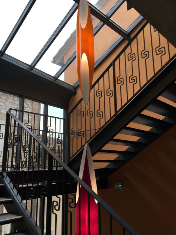 Escalier avec rampe en métal noir, suspensions lumineuses orange et violettes, plafond en verre.