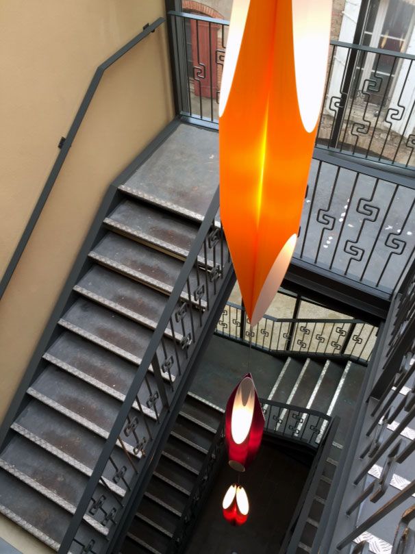 Cage d'escalier avec marches grises, rampes noires et suspensions lumineuses orange.