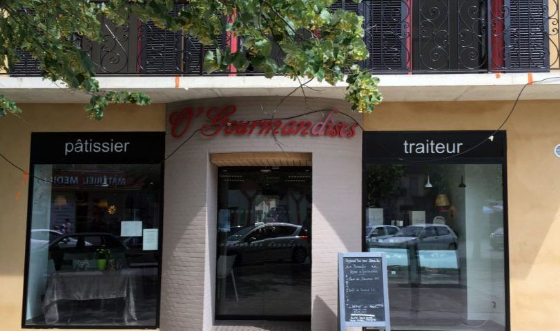 Extérieur de la boulangerie  O' Gourmandises  avec une fenêtre pâtissier  et traiteur, sous un bâtiment à volets.