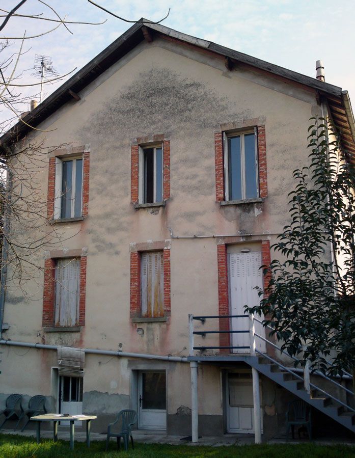 Bâtiment de deux étages, patiné par le temps, avec des encadrements de fenêtres en briques, une porte blanche et un escalier extérieur.
