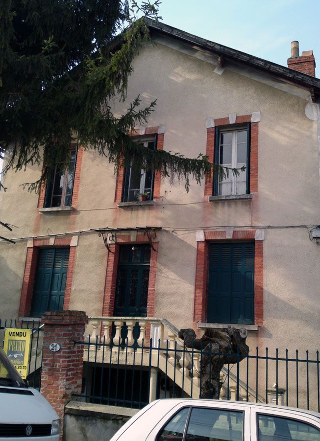 Bâtiment de deux étages avec des encadrements de fenêtres en briques, une clôture en fer forgé et une voiture blanche garée devant.