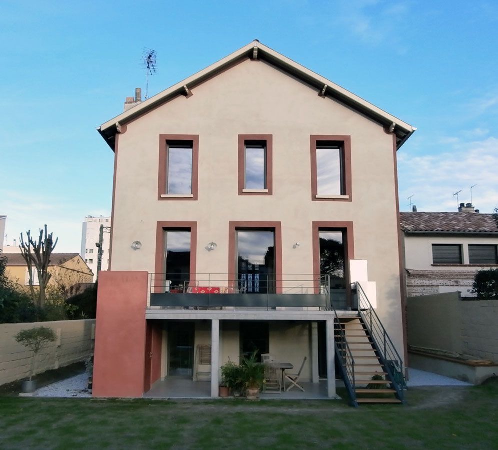 Maison beige à deux étages avec un patio au niveau inférieur, comprenant un escalier et un mur d'accent rouge, sur fond de ciel bleu.