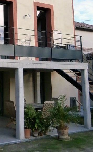 Immeuble avec terrasse extérieure ; plantes en pot et table sous le balcon. Rambarde et escalier noirs.