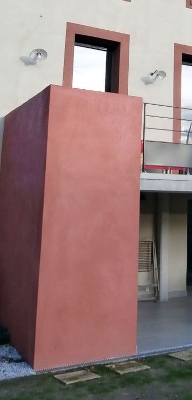 Une haute structure rectangulaire de couleur rouge-brun jouxte un bâtiment doté d'une fenêtre, de deux appliques et d'un patio couvert.