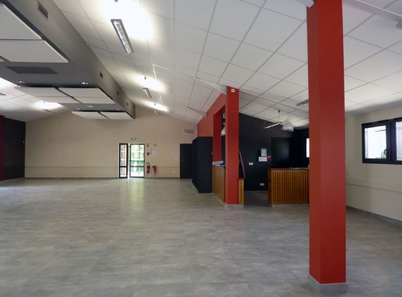 Pièce vide et spacieuse avec sol gris, plafond blanc, piliers rouges et un mur du fond percé d'une porte.