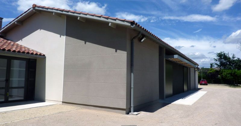 Bâtiment gris au toit de tuiles rouges, situé sur une allée de gravier sous un ciel partiellement nuageux.