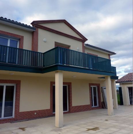 Maison beige avec des touches de brique et un balcon vert, sous un ciel nuageux.