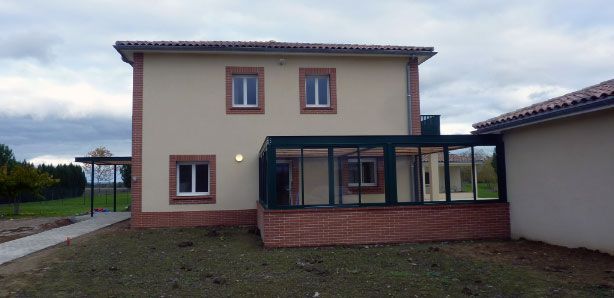Une maison à deux étages avec des accents de briques rouges et un patio vitré, se détachant sur un ciel nuageux.