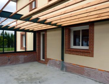 Vue extérieure d'une maison avec une pergola en bois, des éléments en brique et de grandes fenêtres.
