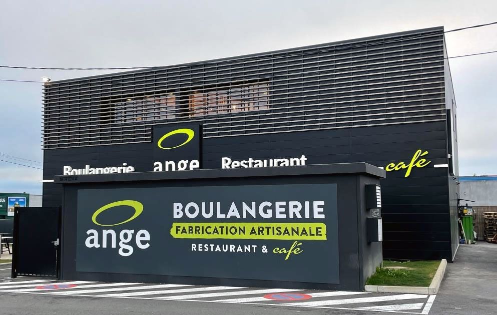 Extérieur de la boulangerie-restaurant Ange. Bâtiment noir avec logos verts et jaunes, inscription Boulangerie Fabrication Artisanale, et un parking.