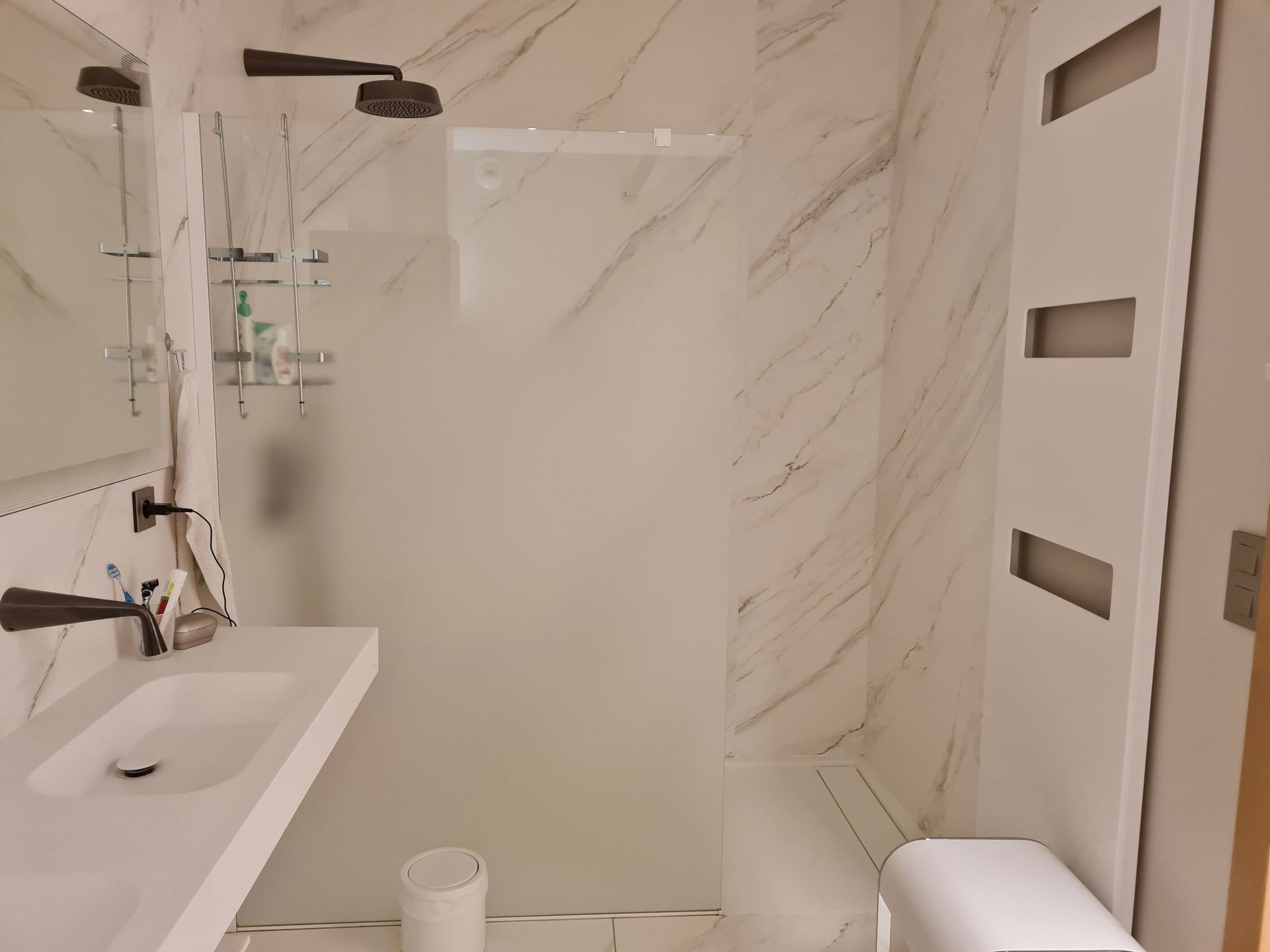 Salle de bains moderne avec carrelage blanc imitation marbre. Comprend une douche, un lavabo et des toilettes avec robinetterie blanche.