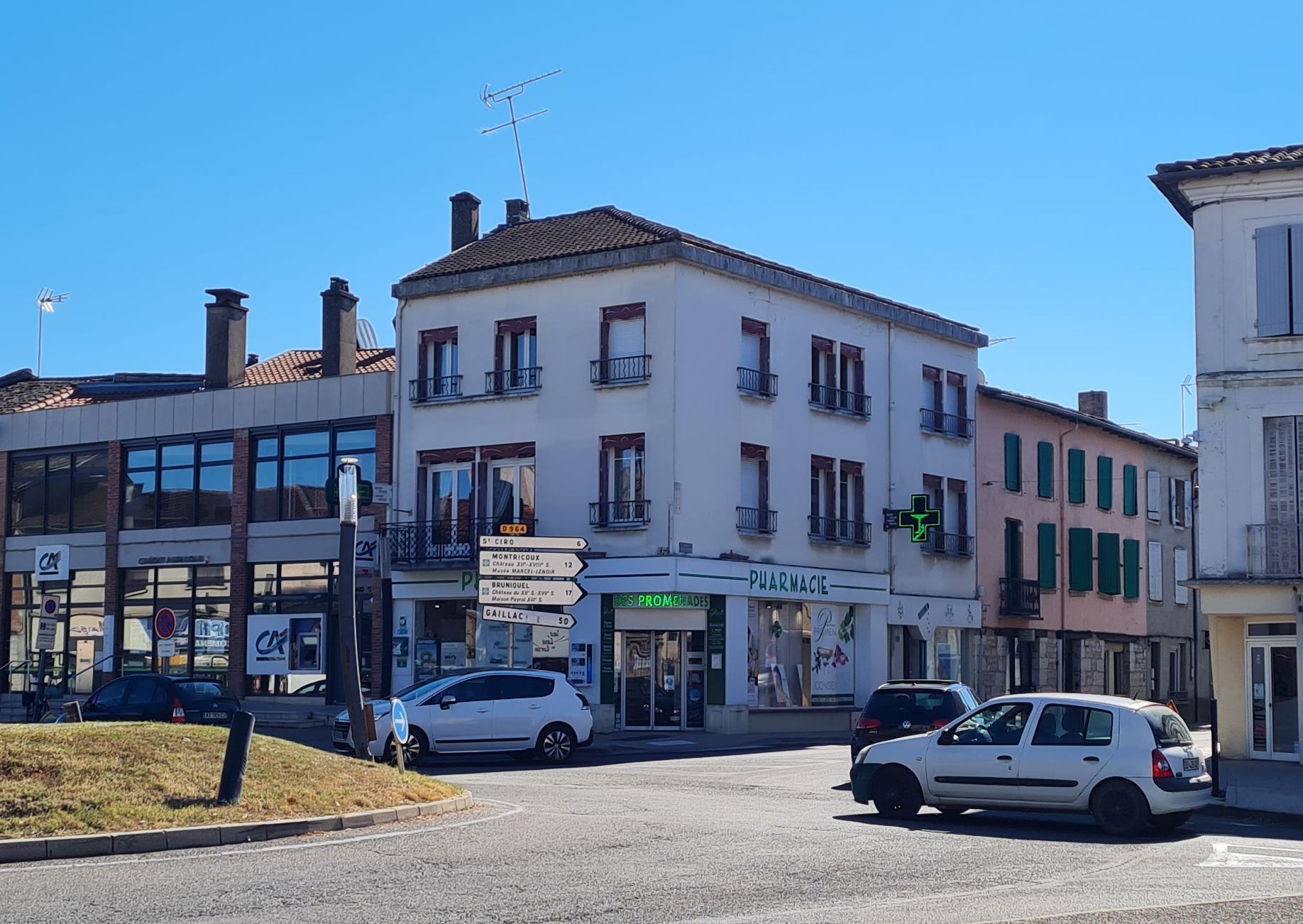 Une pharmacie à un coin de rue, avec des voitures garées, un bâtiment rose et un ciel bleu limpide.