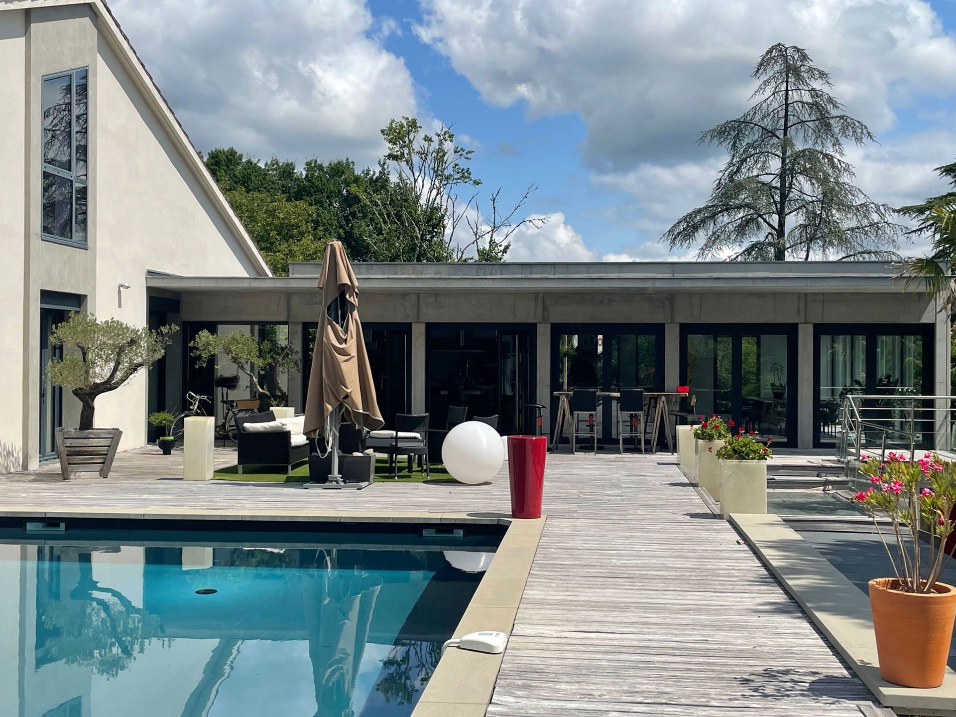 Vue depuis la piscine d'une maison moderne avec portes vitrées, terrasse, arbres et ciel bleu.