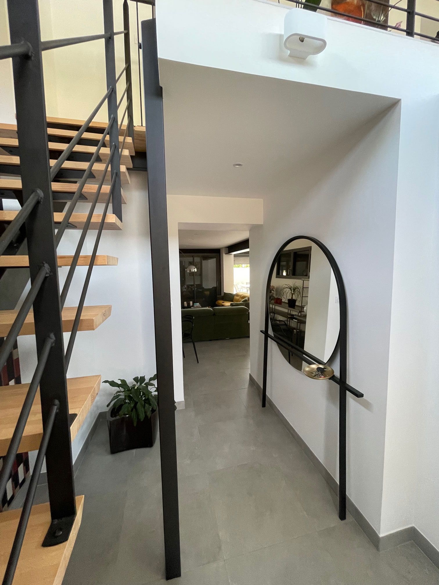 Couloir avec escalier, murs blancs, sol gris, rampe noire et miroir décoratif.