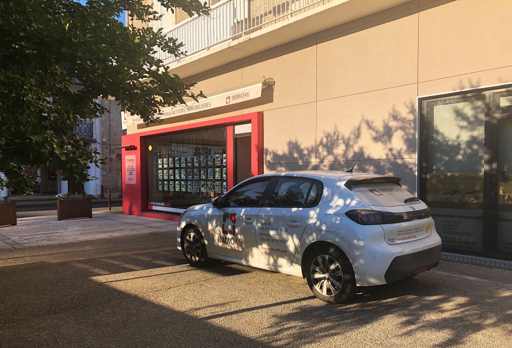 Voiture blanche garée devant un bâtiment à l'entrée rouge. Sol gravillonné, journée ensoleillée.