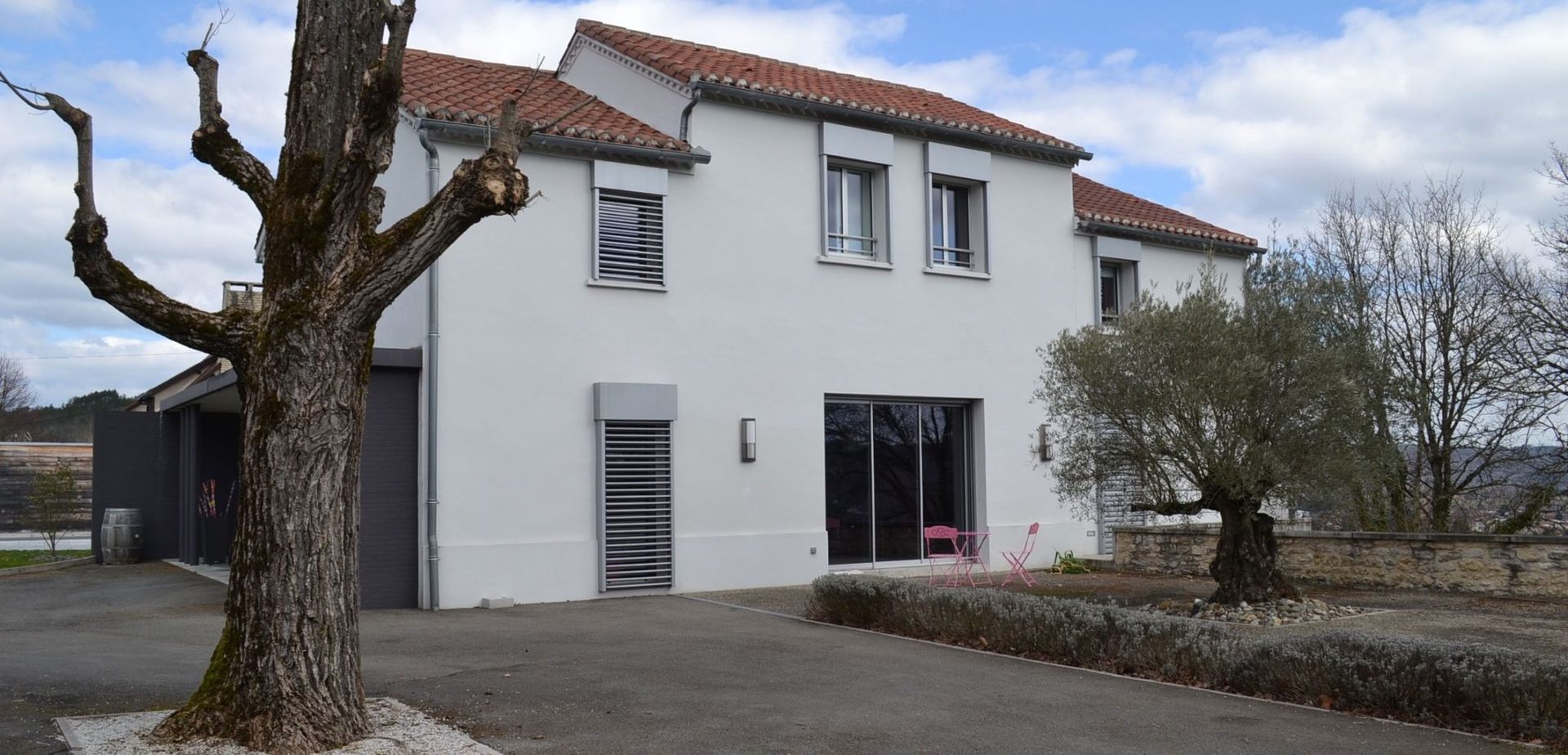 Maison blanche au toit de tuiles rouges, arbres devant. Ciel nuageux.