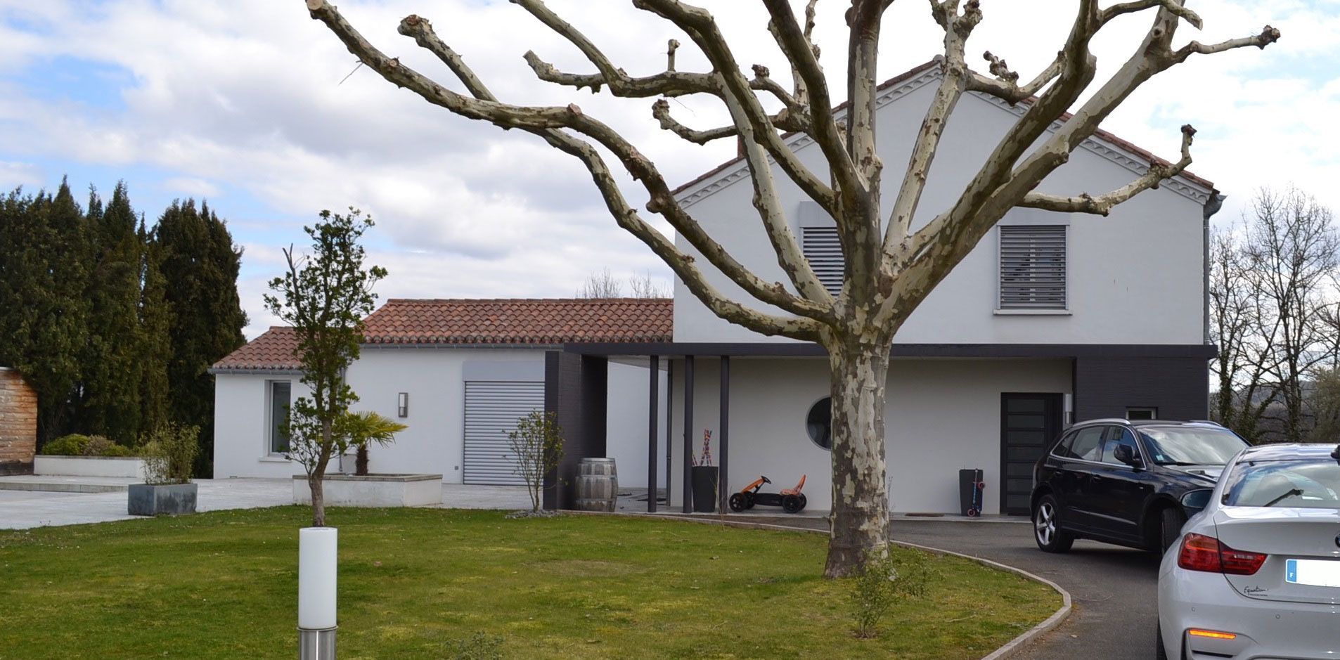 Maison blanche moderne avec un arbre devant et des voitures dans l'allée. Pelouse verte et ciel nuageux.