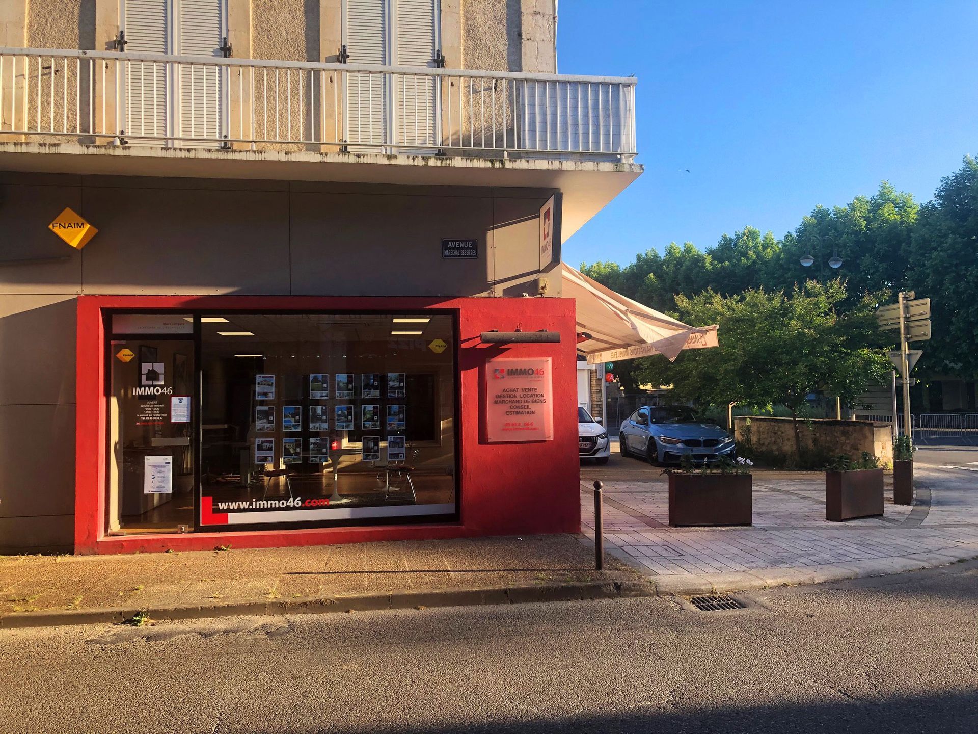 Boutique à l'angle de rue, aux encadrements rouges, vitrine affichant des annonces immobilières. Voitures, arbres et rue en arrière-plan.