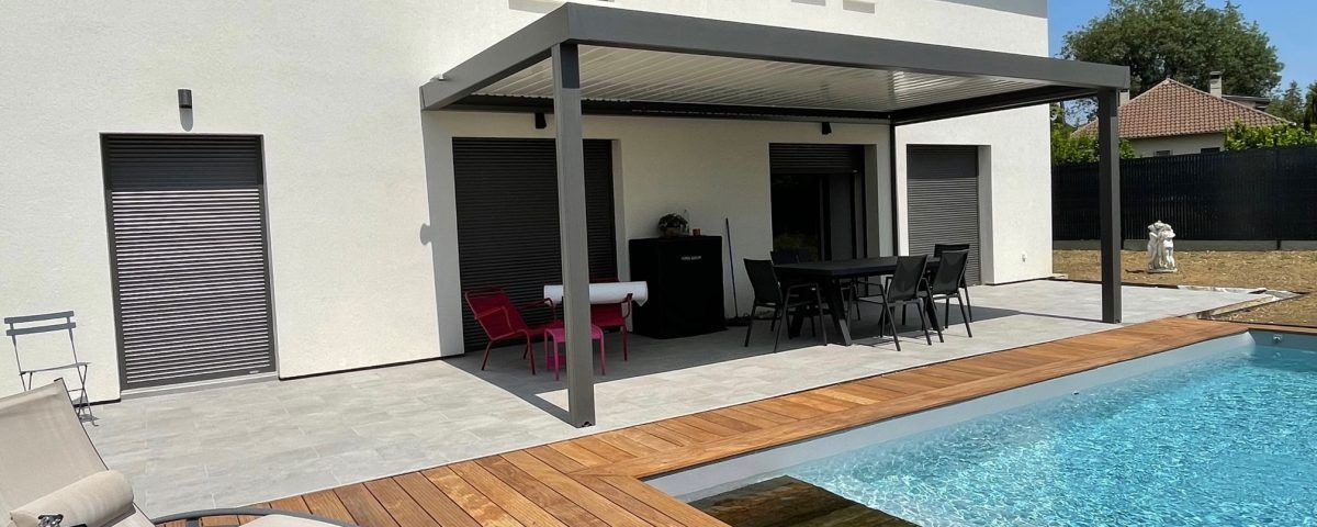 Terrasse avec pergola, table, chaises et piscine. La maison a une façade blanche.