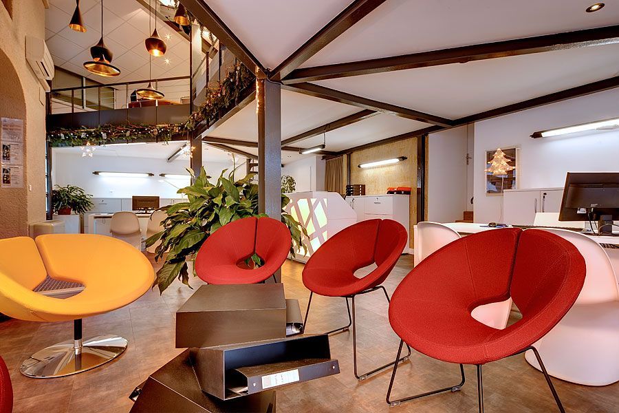 Salle d'attente moderne avec chaises rondes rouges et jaunes, tables en métal et éclairage zénithal.