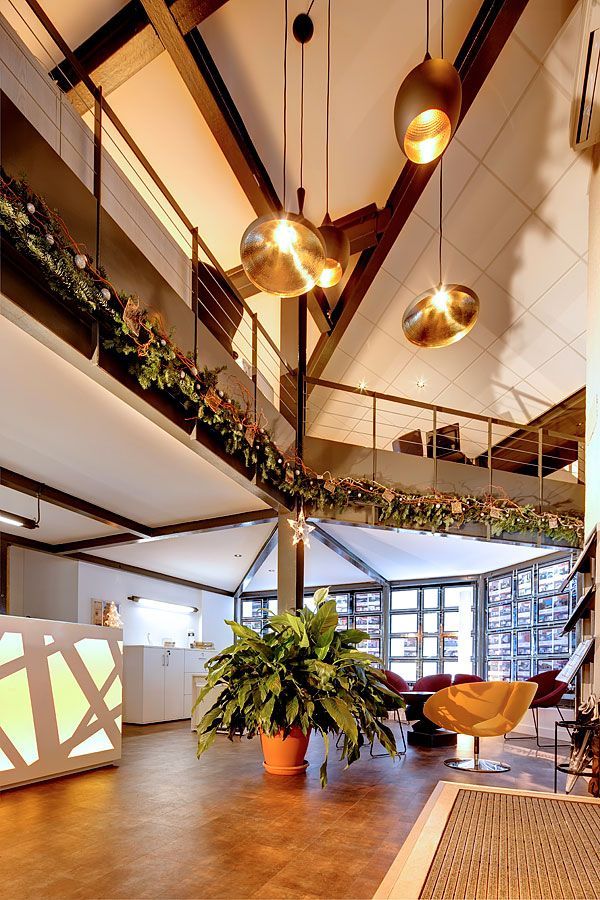 Hall d'entrée avec mezzanine sur deux niveaux. Des suspensions dorées éclairent la pièce, des plantes ornent les balustrades. Une grande plante en pot trône au centre.